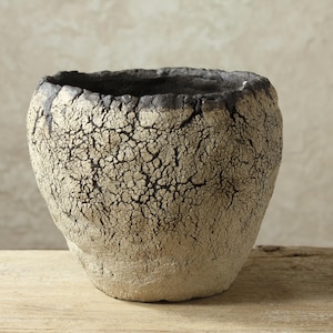 May include: A hand-formed ceramic vessel with a wide, open mouth and a tapered base. The exterior features a heavily textured, crackled off-white finish that transitions into a dark, charred-looking rim with deep fissures and irregular edges.