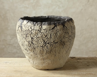 Op de afbeelding: Een handgevormde keramische pot met een wijde opening en een taps toelopende voet. De buitenkant heeft een sterk getextureerde, gebarsten gebroken witte afwerking die overgaat in een donkere, verkoold ogende rand met diepe scheuren.