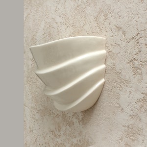 Könnte beinhalten: Weißer Keramik-Wandtopf mit welligem, geriffeltem Design. Der Topf ist an einer strukturierten Wand montiert.