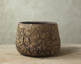 Può includere: Una ciotola in ceramica marrone con una superficie ruvida e screpolata. La ciotola ha un interno scuro e poggia su una superficie di legno. I toni della terra e la trama unica suggeriscono un pezzo fatto a mano o artigianale, adatto all'arredamento.