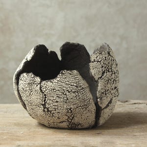 Może przedstawiać: Ręcznie wykonana ceramiczna miska o unikalnej, teksturowanej powierzchni. Misa ma spękaną, kremową powierzchnię zewnętrzną z ciemnoszarym wnętrzem i krawędziami. Organiczny kształt i kontrastujące kolory tworzą uderzający efekt wizualny, do użytku dekoracyjnego.