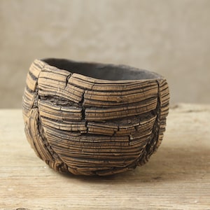 Può includere: Una piccola ciotola in ceramica rotonda con un esterno strutturato simile alla corteccia in tonalità marrone e marrone chiaro. La superficie presenta profondi solchi orizzontali e crepe verticali, mentre l'interno è di un liscio grigio scuro.