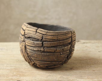 May include: A small, round ceramic bowl with a textured, bark-like exterior in shades of brown and tan. The surface features deep, horizontal grooves and vertical cracks, while the interior of the bowl is a smooth, dark charcoal gray color.