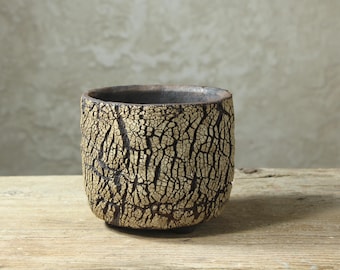 Könnte beinhalten: Ein kleiner, zylindrischer Keramikbecher mit einer dunkel anthrazitfarbenen Basisschicht, die von einer dicken, cremefarbenen Glasur mit einer ausgeprägten, tief rissigen Textur bedeckt ist. Das Innere des Bechers ist dunkel und die Außenseite ist rau.
