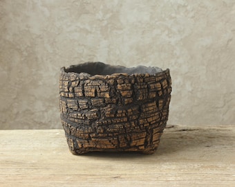 May include: A round ceramic planter with a textured exterior that mimics the appearance of dark brown tree bark. The vessel features a deep, hollow interior with a dark gray finish and an uneven, organic rim, showcasing a rugged, weathered aesthetic.