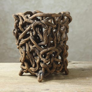 May include: A cylindrical vessel constructed from a dense, interconnected network of brown, textured, branch-like forms. The structure features an open-work design with irregular gaps throughout, supported by several small, protruding feet at the base.