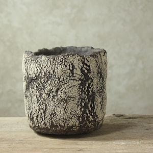 May include: A cylindrical ceramic vessel featuring a dark brown base color with a thick, heavily crackled off-white glaze overlay. The rim is uneven and textured, and the exterior surface displays a deep, web-like pattern of fissures.