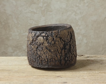 May include: A small, cylindrical ceramic cup with a dark brown, heavily textured exterior featuring a cracked, weathered surface pattern. The rim is uneven and thick, with subtle metallic gold highlights accentuating the deep fissures in the clay.