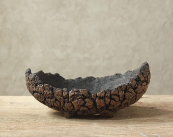 May include: A handcrafted, textured ceramic bowl with a dark brown exterior and a gray interior. The bowl has a unique, organic shape and sits on three small feet. The surface has a rough, bark-like texture, perfect for displaying small items.