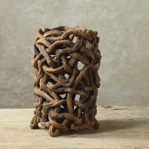May include: A cylindrical vase constructed from a dense, intricate network of brown, textured, branch-like elements. The structure features an open, lattice-style design with numerous gaps between the intertwined, gnarled wooden-looking segments.
