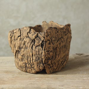Può includere: Un vaso quadrato fatto a mano con una superficie ruvida e strutturata. Il vaso è realizzato in un materiale marrone chiaro con fessure marroni scure, che gli conferiscono un aspetto naturale e invecchiato. L'apertura è irregolare, aggiungendo al suo design unico.