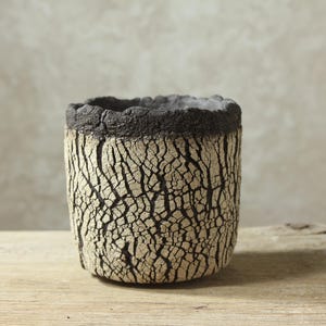 Peut inclure: Un récipient cylindrique en céramique présentant une surface extérieure crème fortement texturée et craquelée avec des fissures noires profondes. Le bord supérieur est épais, irrégulier et de couleur anthracite, contrastant avec le motif.