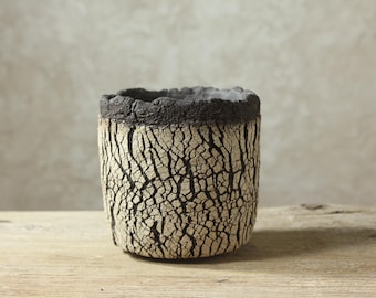 Può includere: Un vaso cilindrico in ceramica caratterizzato da una superficie esterna color crema, fortemente strutturata e screpolata, con profonde fessure nere. Il bordo superiore è spesso, irregolare e di colore antracite, in contrasto con il motivo.