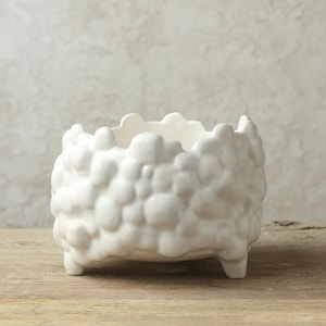 Op de afbeelding: Witte keramische plantenbak met een getextureerd, hobbelig oppervlak en drie korte pootjes. De plantenbak is komvormig en heeft een afgeronde, organische vorm.