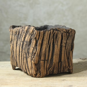 May include: A square ceramic planter with a textured exterior that mimics the appearance of tree bark. The surface features deep vertical grooves and ridges in shades of brown and dark charcoal, with a dark gray interior rim.