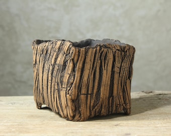 May include: A square ceramic planter with a textured exterior that mimics the appearance of tree bark. The surface features deep vertical grooves and ridges in shades of brown and dark charcoal, with a dark grey interior rim.