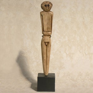 May include: A carved wooden figurine of a person with a simplified face and body. The figurine is standing on a black base.