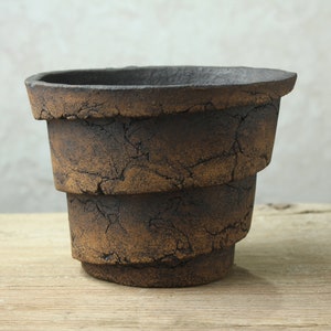May include: A brown ceramic planter with a textured, cracked surface. The planter has a stepped design and a rounded base.