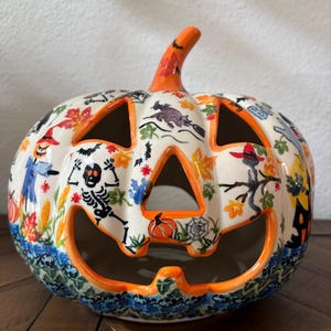 May include: A ceramic jack-o'-lantern featuring a cream-colored base with hand-painted Halloween motifs including skeletons, witches, bats, pumpkins, and autumn leaves. The pumpkin has cut-out eyes, nose, and mouth with bright orange trim.