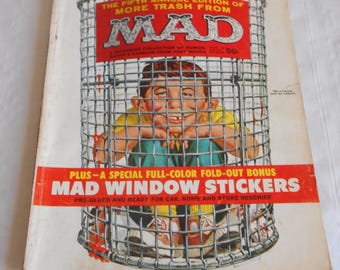 May include: A vintage Mad Magazine cover featuring a boy trapped in a wire trash can. The cover text reads "The Fifth Annual Edition of More Trash From Mad". The cover also includes a bonus offer for "Mad Window Stickers".