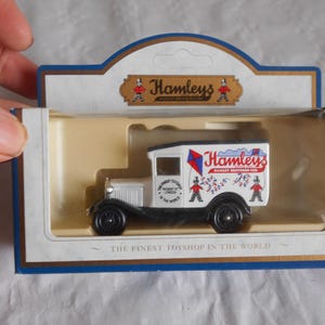 May include: A white toy van with a red and blue Hamleys logo and the text "Hamleys,  Hamleys Brothers Ltd."  The van is in a clear plastic display case with a white background and the text "The Finest Toyshop in the World".