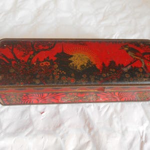 Op de afbeelding: Rechthoekige rode en zwarte vintage blikken doos met een Aziatisch geïnspireerd ontwerp. De deksel toont een zonsondergang met een pagode, bomen en een vogel. De zijkanten hebben bloemmotieven. De blikken doos is ongeveer 25 cm lang.