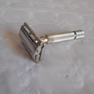 Op de afbeelding: Een vintage, verchroomd veiligheidsscheermes met een getextureerde handgreep. De scheerkop is rechthoekig met een scharnierend ontwerp. De handgreep heeft een gekarteld patroon voor grip. Het scheermes is ontworpen om te scheren.