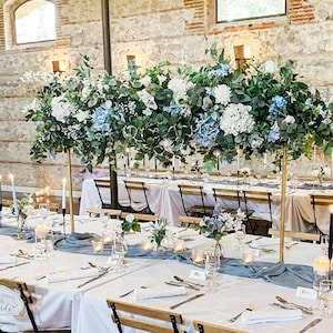 May include: A wedding reception table setting with white tablecloths, blue table runners, and black chairs. The table is decorated with white and blue flowers, candles, and greenery. The table is set with white napkins, cutlery, and glasses.