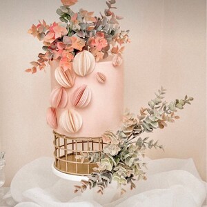 May include: A pink cake decorated with paper flowers and greenery, sitting on a gold wire cake stand.