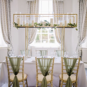 May include: A wedding reception table setting with three chairs draped in sage green fabric. The chairs are arranged in a row facing a window with white curtains. A gold metal frame with clear glass cylinders holds white candles and greenery.