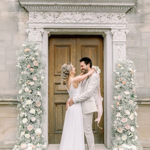Könnte beinhalten: Eine Braut und ein Bräutigam stehen vor einer mit weißen und rosa Blumen geschmückten Tür. Der Bräutigam trägt einen hellbraunen Anzug und die Braut ein weißes Kleid. Sie lächeln sich an und schauen sich an.