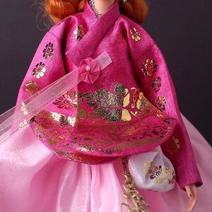 Pode incluir: Uma boneca vestindo um hanbok coreano tradicional. O traje rosa brilhante apresenta bordados florais dourados e uma saia rosa combinando. Uma pequena bolsa branca com detalhes dourados pende do pulso da boneca. O hanbok &eacute; adornado com uma fita decorativa e uma p&eacute;rola.