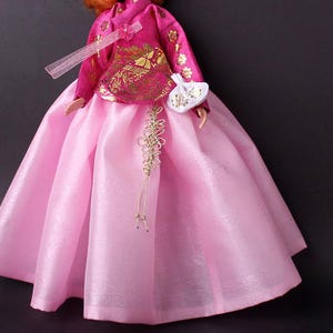 Pode incluir: Boneca vestida com um tradicional Hanbok coreano. O vestido apresenta uma saia rosa vibrante e um top magenta com bordados florais dourados. Uma pequena bolsa branca e detalhes dourados completam o conjunto. A boneca tem cabelo ruivo.
