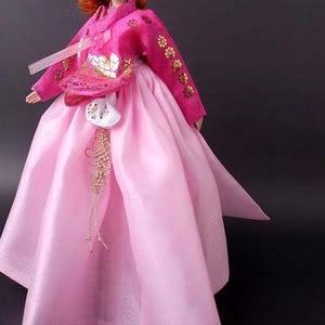 Pode incluir: Boneca vestindo um tradicional vestido Hanbok coreano. O vestido &eacute; rosa vibrante com bordados florais dourados nas mangas e no corpete. Uma bolsa branca com detalhes dourados pende da cintura, e a saia &eacute; rosa claro e fluida.