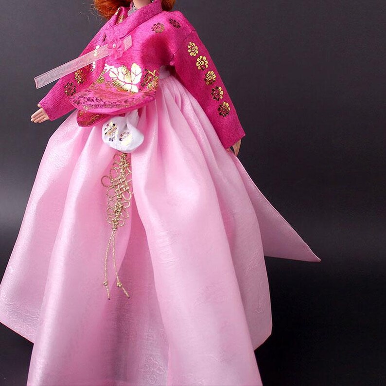 Pode incluir: Boneca vestindo um tradicional vestido Hanbok coreano. O vestido &eacute; rosa vibrante com bordados florais dourados nas mangas e no corpete. Uma bolsa branca com detalhes dourados pende da cintura, e a saia &eacute; rosa claro e fluida.
