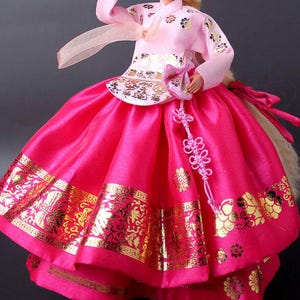 Puede incluir: Mu&ntilde;eca con un vestido Hanbok coreano tradicional. El vestido presenta una parte superior rosa con detalles florales dorados y una falda fucsia vibrante con ribete dorado. Un accesorio rosa cuelga de la cintura.