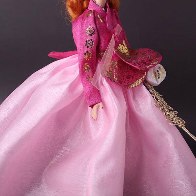 Pode incluir: Boneca vestida com um hanbok coreano tradicional. O vestido rosa vibrante apresenta detalhes florais dourados na jaqueta e uma saia fluida. A boneca de cabelo ruivo est&aacute; em um fundo escuro.