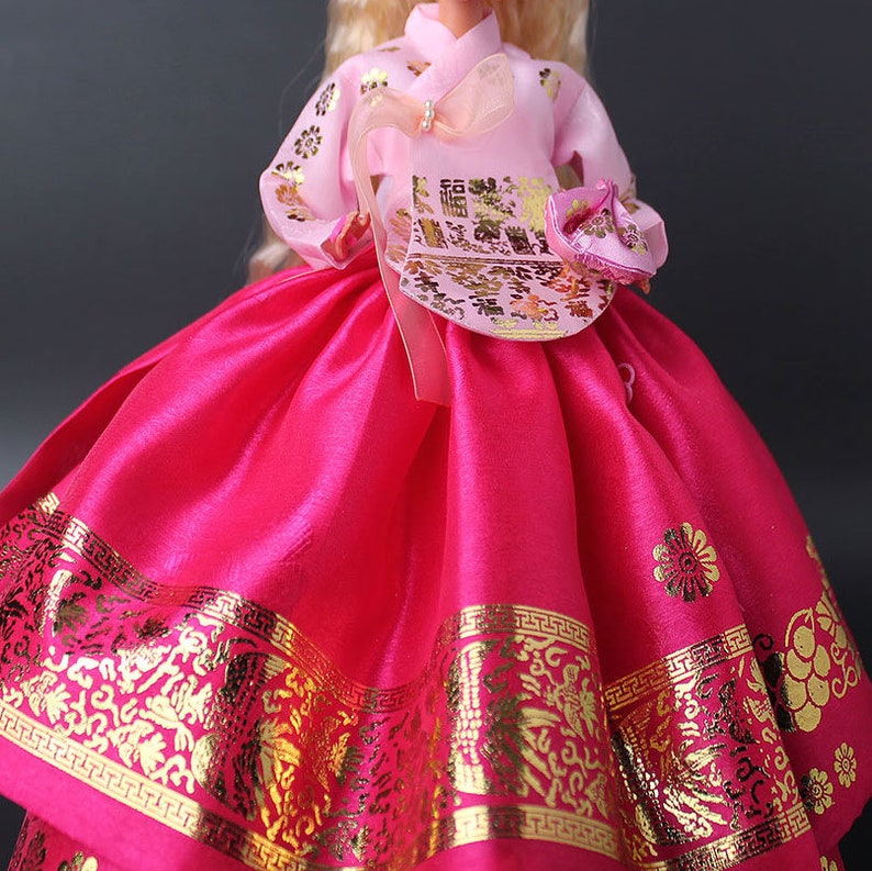 Puede incluir: Mu&ntilde;eca vestida con un tradicional vestido Hanbok coreano. El vestido es de color rosa vibrante con patrones florales y geom&eacute;tricos dorados. La parte superior es de color rosa claro con detalles dorados, e incluye un peque&ntilde;o bolso a juego.