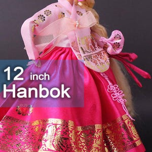 Puede incluir: Vestido de mu&ntilde;eca Hanbok de 30 cm en tonos rosa y rojo, con detalles dorados. La prenda tradicional coreana presenta intrincados estampados florales y un lazo decorativo. El texto "12 inch Hanbok" aparece en la imagen.