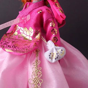 Pode incluir: Um hanbok coreano tradicional em tons de rosa, adornado com padr&otilde;es florais dourados. A jaqueta apresenta um la&ccedil;o e uma pequena bolsa branca com detalhes dourados. A saia &eacute; de um rosa mais claro, criando um efeito em camadas.