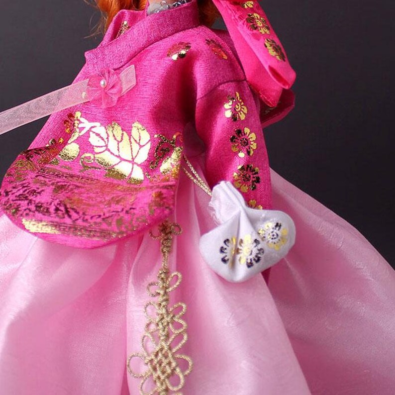 Pode incluir: Um hanbok coreano tradicional em tons de rosa, adornado com padr&otilde;es florais dourados. A jaqueta apresenta um la&ccedil;o e uma pequena bolsa branca com detalhes dourados. A saia &eacute; de um rosa mais claro, criando um efeito em camadas.