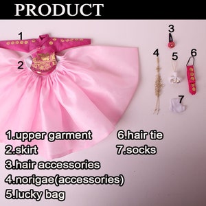 Pode incluir: Um traje tradicional de boneca coreana, com uma parte superior e saia rosa, com detalhes dourados. Os acess&oacute;rios incluem grampos de cabelo, um saco da sorte e meias. O traje &eacute; para uma boneca.