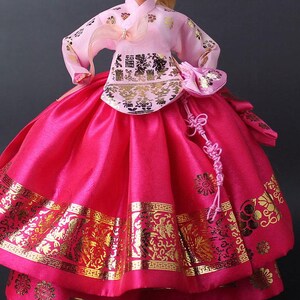 Puede incluir: Mu&ntilde;eca vestida con un tradicional vestido Hanbok coreano. El vestido presenta una parte superior rosa con detalles dorados y una falda fucsia vibrante. La falda tiene patrones dorados en el dobladillo. Un peque&ntilde;o bolso rosa y un nudo decorativo completan el conjunto.