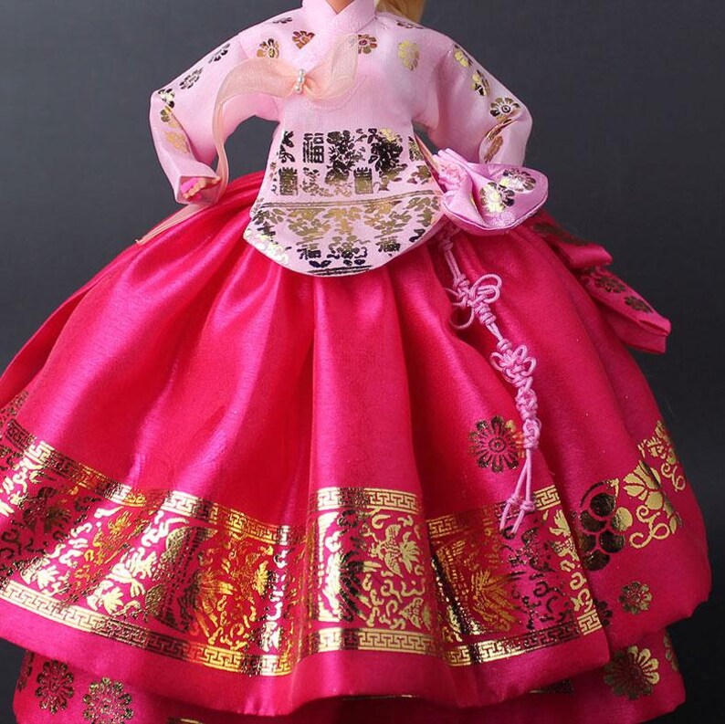 Puede incluir: Mu&ntilde;eca vestida con un tradicional vestido Hanbok coreano. El vestido presenta una parte superior rosa con detalles dorados y una falda fucsia vibrante. La falda tiene patrones dorados en el dobladillo. Un peque&ntilde;o bolso rosa y un nudo decorativo completan el conjunto.