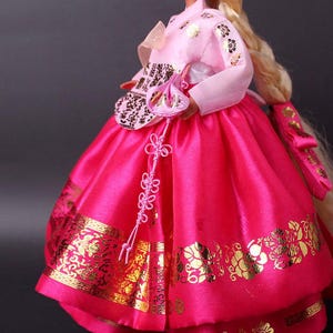 Puede incluir: Una mu&ntilde;eca con un vestido Hanbok coreano tradicional. El vestido presenta una falda rosa brillante con patrones florales dorados y una parte superior rosa claro con detalles dorados. Un peque&ntilde;o bolso a juego y un nudo decorativo completan el conjunto.