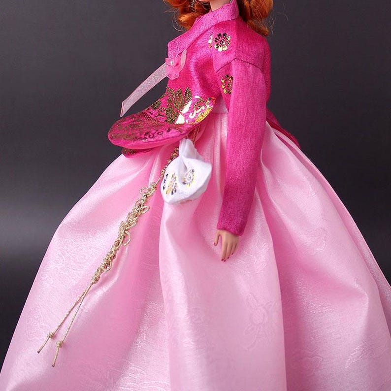 Pode incluir: Uma boneca vestindo um tradicional vestido Hanbok coreano. O vestido &eacute; rosa vibrante, complementado por uma jaqueta combinando adornada com bordados florais dourados. Uma pequena bolsa branca com detalhes florais pende de um cord&atilde;o dourado.