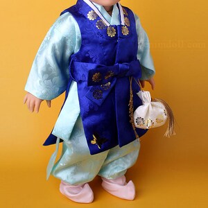 Puede incluir: Una muñeca vestida con un traje tradicional coreano. El conjunto presenta un chaleco azul vibrante con detalles florales dorados sobre una camisa y pantalones azul claro. Un lazo azul a juego y una pequeña bolsa blanca con diseños florales dorados completan el look. La muñeca también lleva zapatos rosas.