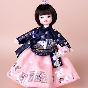 May include: A doll dressed in a traditional Korean Hanbok. The outfit includes a navy blue jacket with silver floral details and a pink skirt with silver patterns. The doll has dark hair and a small pink headpiece.