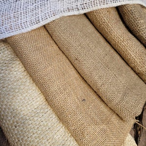 May include: Close-up of various rolls of burlap fabric in shades of tan and beige. A white, open-weave ribbon is draped over the top. The coarse texture of the burlap is visible, suggesting a rustic or natural aesthetic, suitable for crafts or decor.