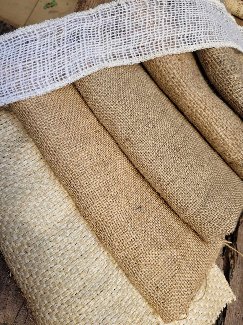 May include: Close-up of various rolls of burlap fabric in shades of tan and beige. A white, open-weave ribbon is draped over the top. The coarse texture of the burlap is visible, suggesting a rustic or natural aesthetic, suitable for crafts or decor.